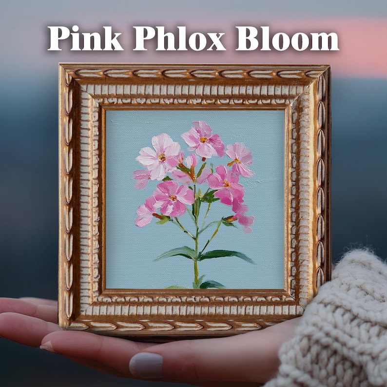 May include: A square framed oil painting featuring a cluster of pink phlox flowers against a light blue background. The ornate gold frame has a textured, repeating leaf pattern. The text "Pink Phlox Bloom" is printed in white above the frame.