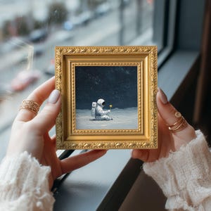 May include: A small, square, gold-framed painting depicts an astronaut seated on a lunar surface, holding a yellow flower. The artwork is held by a person, showcasing the painting's intricate details and the gold frame's ornate design. The background is a dark, starry sky.