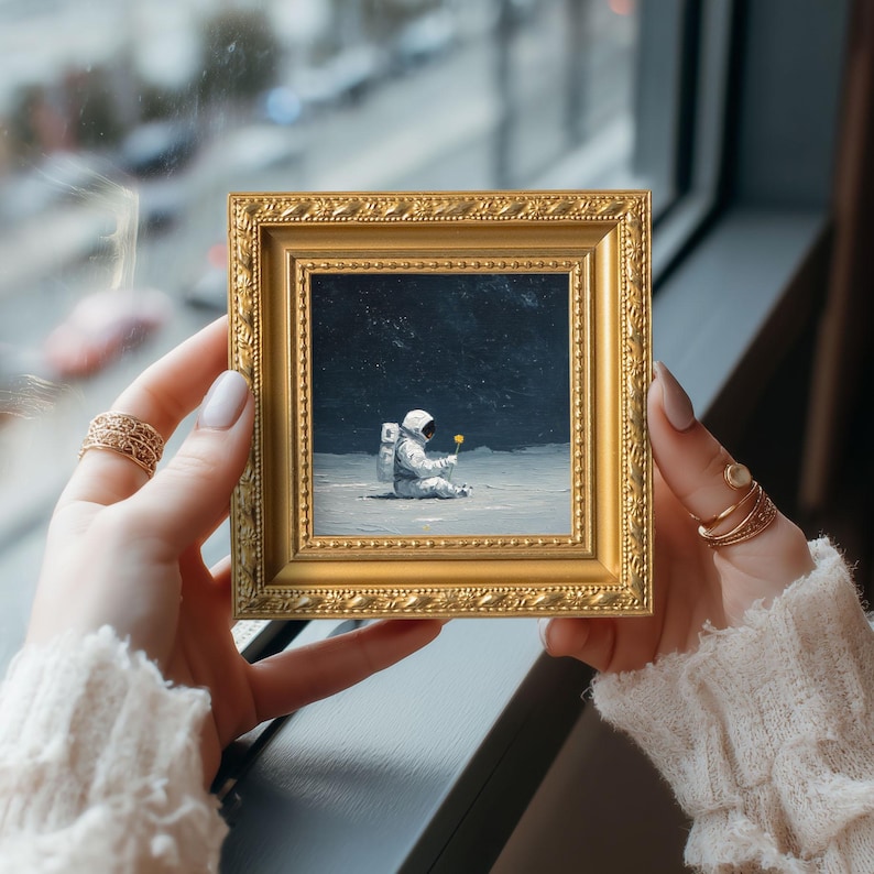 May include: A small, square, gold-framed painting depicts an astronaut seated on a lunar surface, holding a yellow flower. The artwork is held by a person, showcasing the painting's intricate details and the gold frame's ornate design. The background is a dark, starry sky.