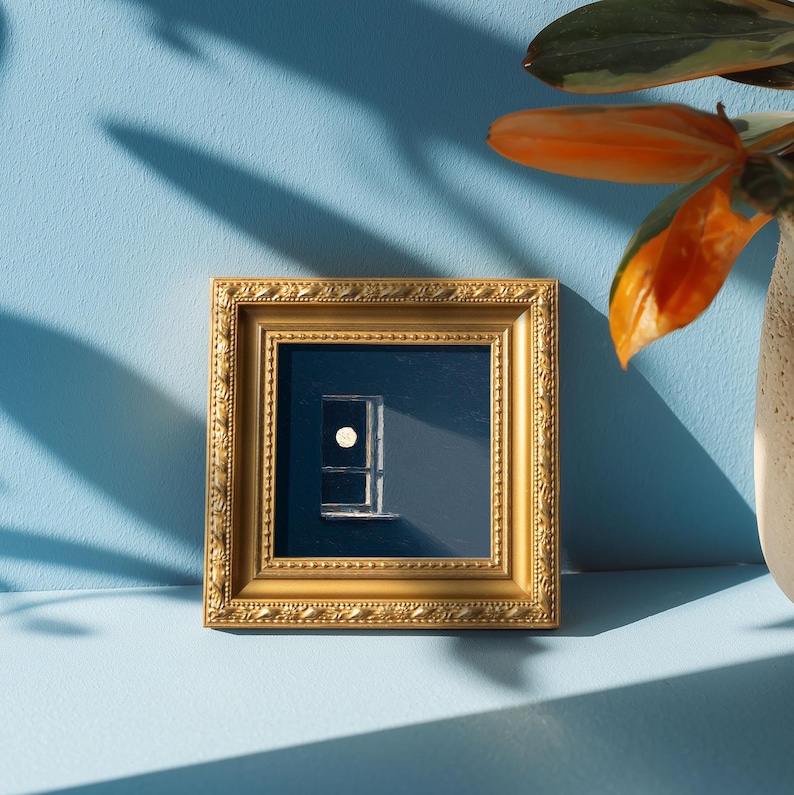 May include: A small framed artwork featuring a minimalist design of a window with a circular element. The frame is gold-colored with ornate detailing. The artwork is set against a dark blue background, with a light blue wall in the background.