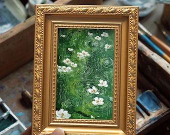 May include: A small, framed painting featuring white flowers floating on a green pond with concentric circles. The painting is in a gold ornate frame. The artwork is held by a person's hand, with art supplies visible in the background.