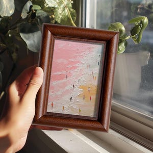 May include: A framed miniature painting depicting a beach scene with tiny figures. The artwork features a pink and white sky, a sandy shore, and a textured, impasto style. The frame is made of dark brown wood.