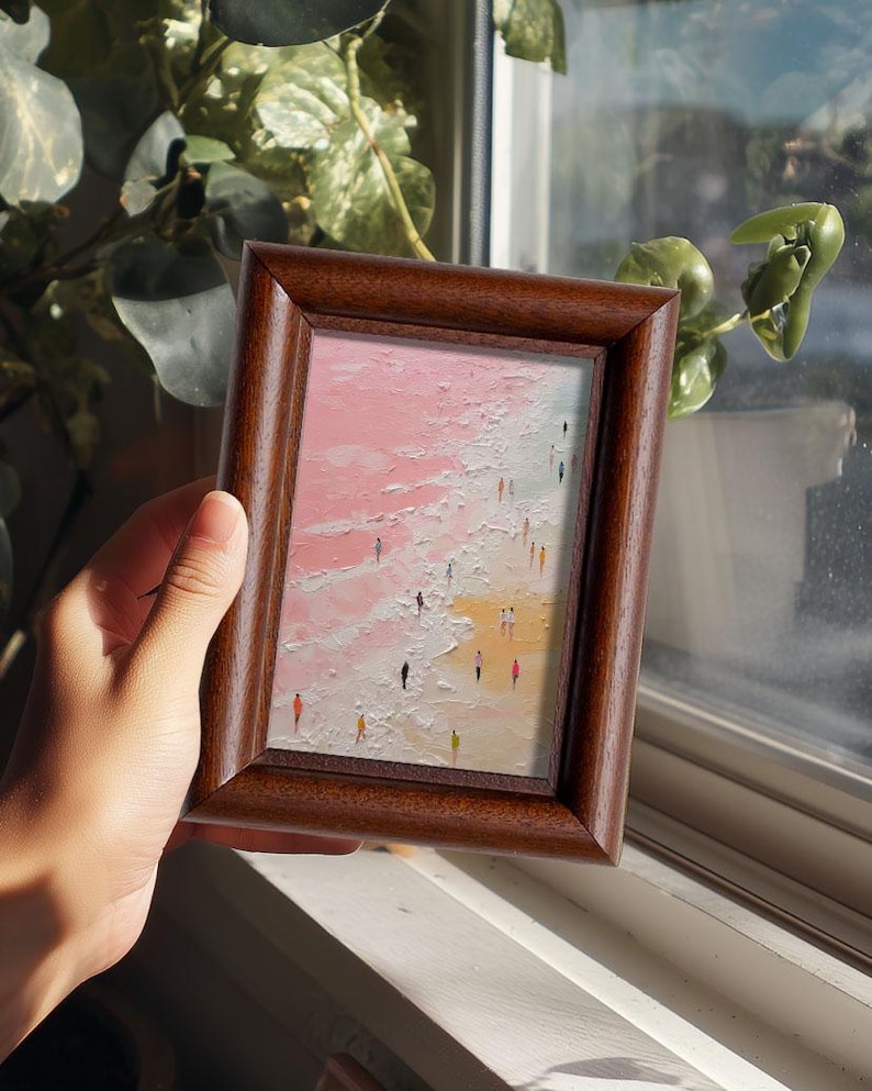 May include: A framed miniature painting depicting a beach scene with tiny figures. The artwork features a pink and white sky, a sandy shore, and a textured, impasto style. The frame is made of dark brown wood.