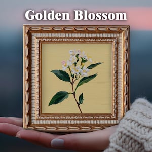 May include: A small square painting in a decorative gold frame featuring a white flower with green leaves on a pale yellow background. The text Golden Blossom is printed in white above the frame. The artwork displays visible brushstrokes.