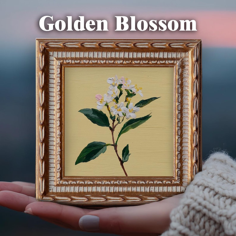 May include: A small square painting in a decorative gold frame featuring a white flower with green leaves on a pale yellow background. The text Golden Blossom is printed in white above the frame. The artwork displays visible brushstrokes.