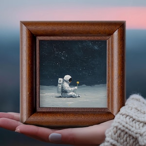 May include: A framed painting depicts an astronaut seated on a lunar surface, holding a yellow flower. The artwork is set against a dark, starry sky. The frame is made of dark wood.