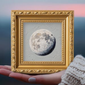 May include: A small square oil painting of a textured, cratered moon in shades of white, gray, and dark blue, set against a light gray background. The artwork is encased in an ornate, metallic gold frame with a beaded and embossed border design.
