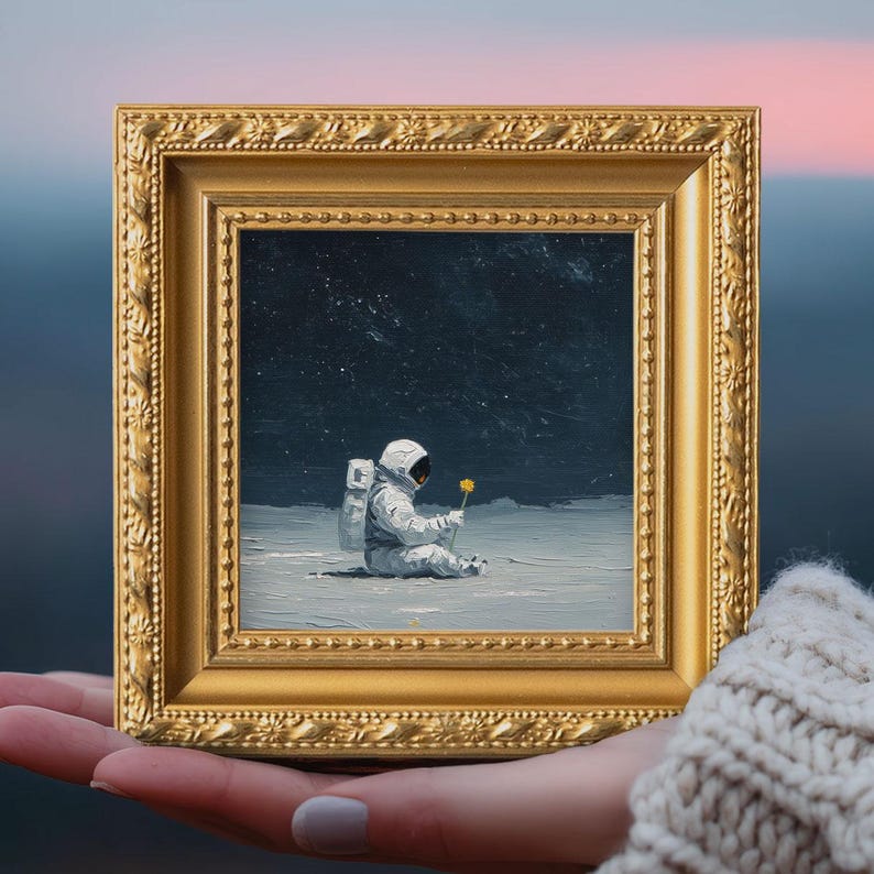 May include: A framed painting depicts an astronaut seated on a lunar surface, holding a yellow flower. The artwork is set against a dark, starry sky. The gold frame adds a classic touch to the space-themed art.