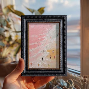 May include: A framed miniature painting depicts a beach scene with tiny figures. The artwork features a pink and white sky and water, with a sandy shore. The black frame has an ornate, textured design. The painting is held in a hand.