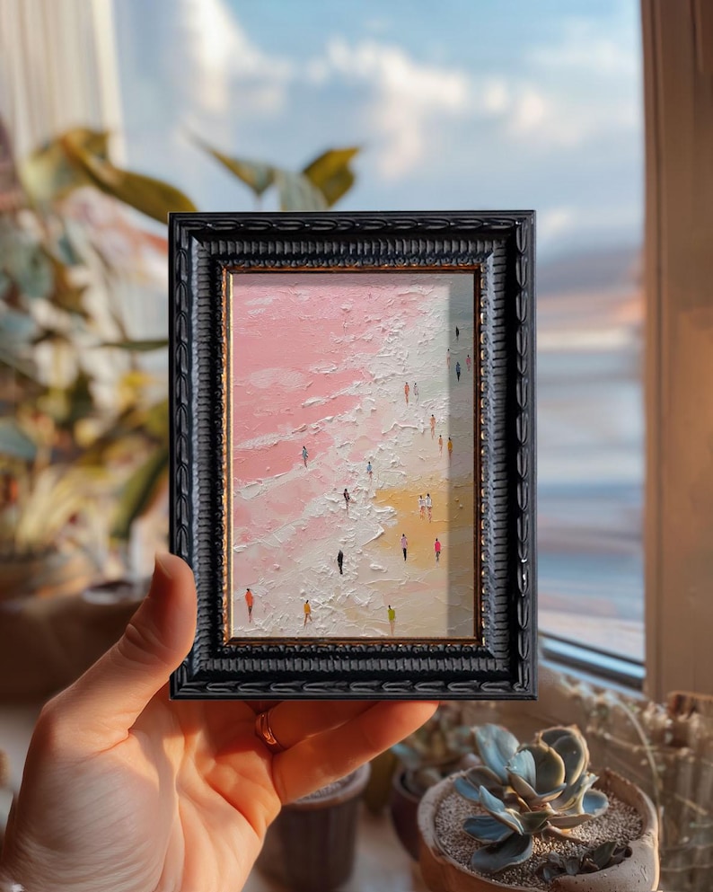 May include: A framed miniature painting depicts a beach scene with tiny figures. The artwork features a pink and white sky and water, with a sandy shore. The black frame has an ornate, textured design. The painting is held in a hand.