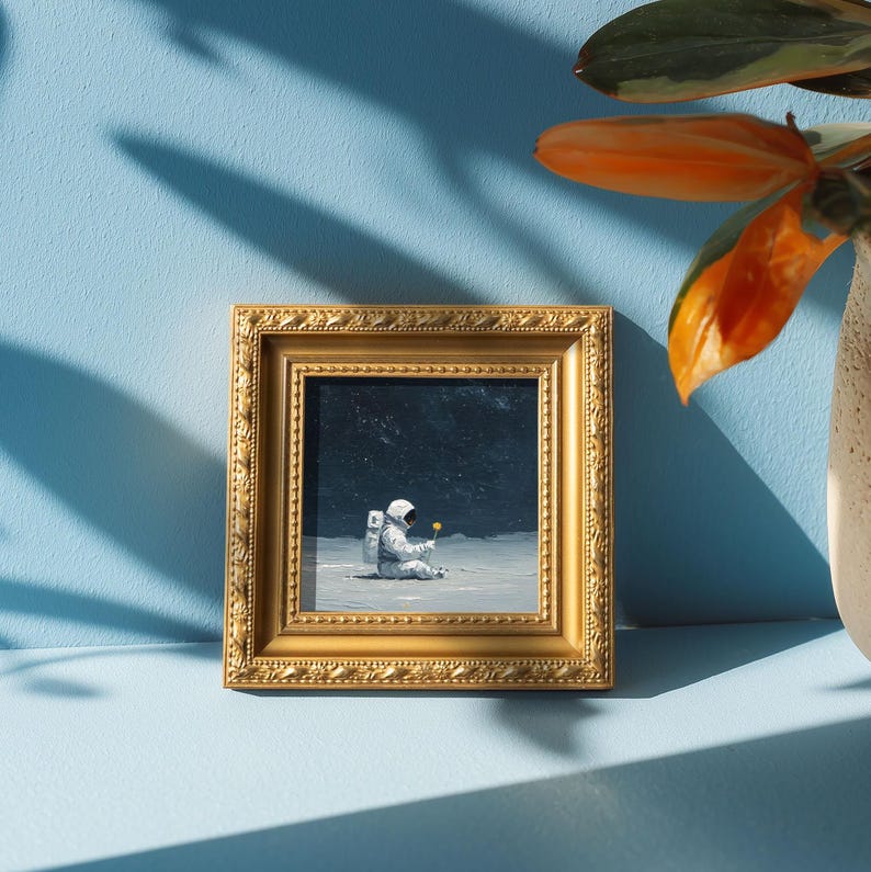 May include: A framed painting depicts an astronaut seated on a lunar surface, holding a yellow flower. The artwork is encased in a gold-colored, ornate frame. The background is a light blue surface with shadows.