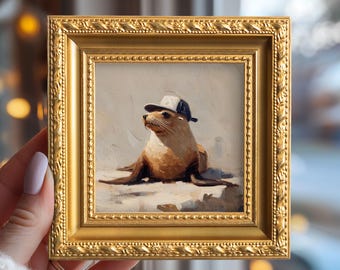 May include: A framed painting of a sea lion wearing a baseball cap. The artwork is in a gold ornate frame. The sea lion is brown and tan, and the cap is white and navy blue. The background is a light, neutral color.