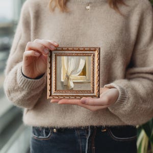May include: Small framed painting depicting a window with a white curtain. The painting has a gold frame with a white inner border. The artwork is held by a person wearing a beige sweater and blue jeans.