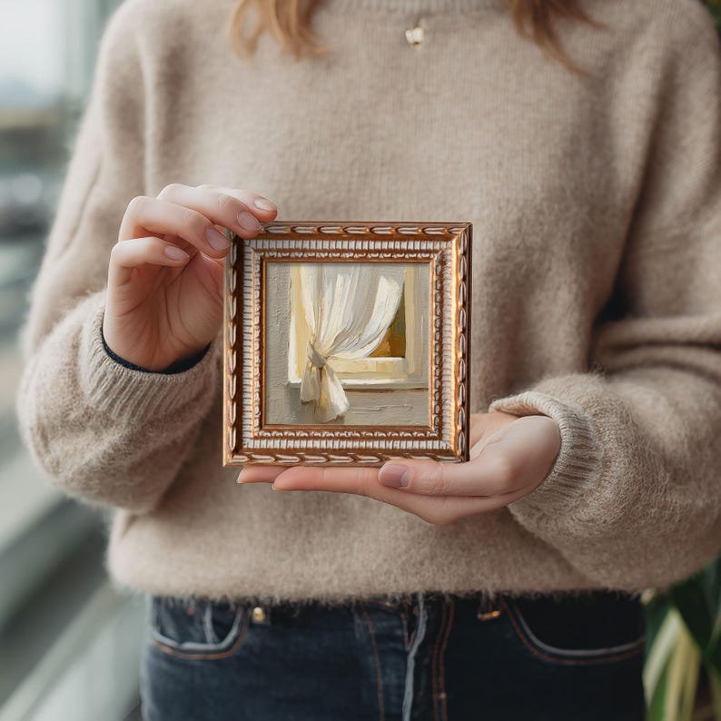 May include: Small framed painting depicting a window with a white curtain. The painting has a gold frame with a white inner border. The artwork is held by a person wearing a beige sweater and blue jeans.