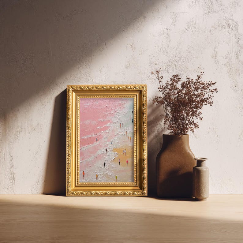 May include: A framed painting with a gold ornate frame depicts a beach scene with figures. The artwork features a pink and white sky, a sandy shore, and tiny figures. A brown vase with dried flowers and a small vase are next to the painting.