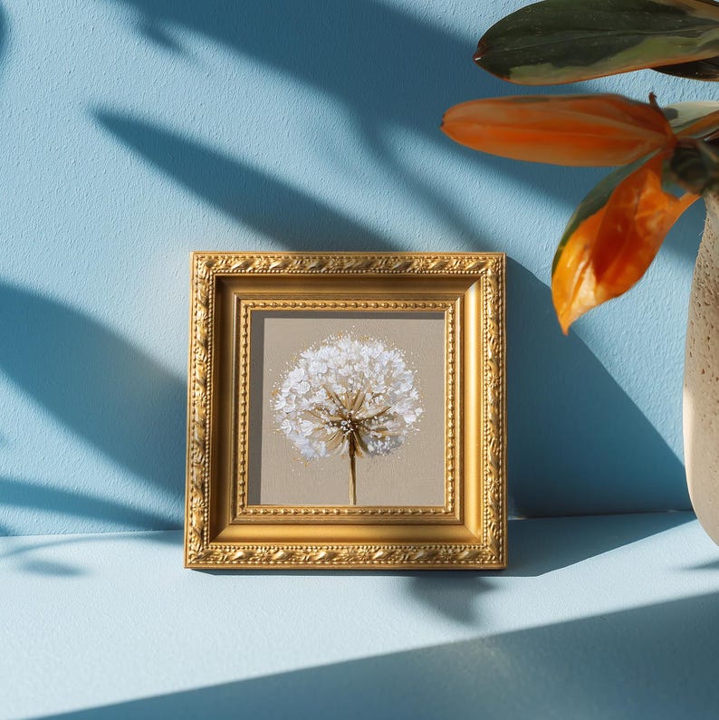 May include: A square wall art piece featuring a white dandelion seed head illustration on a beige background, encased in an ornate, metallic gold frame with intricate embossed detailing along the edges of the border.