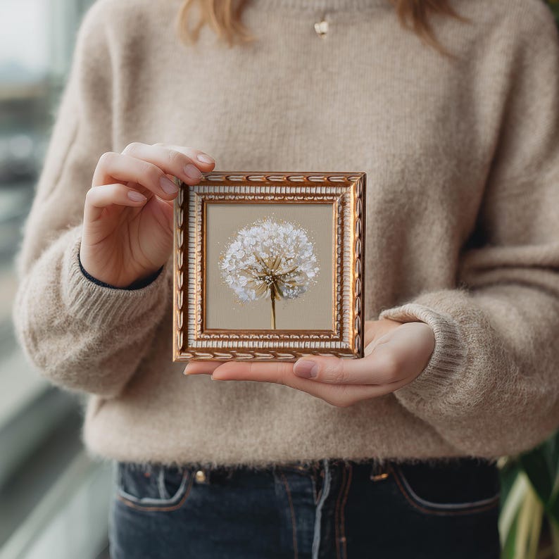 May include: A square wall art piece featuring a white dandelion seed head illustration on a beige background, encased in an ornate, metallic gold-toned frame with a textured, repeating leaf-like pattern along the outer border and inner edge.