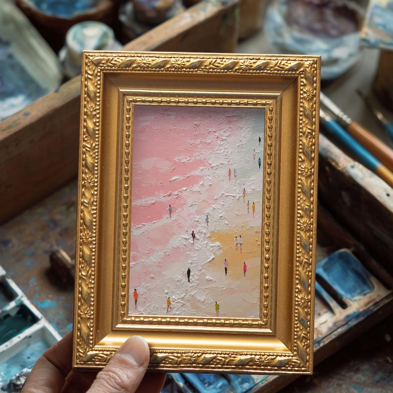 May include: A framed miniature painting depicts a beach scene with figures. The artwork features a gold ornate frame. The painting shows a pink sky, white waves, and a yellow beach with tiny figures in various colors.