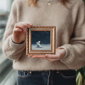 May include: A small framed painting depicts an astronaut seated on a lunar surface, holding a yellow flower. The artwork is set against a dark, starry background. The frame is gold with a detailed border.