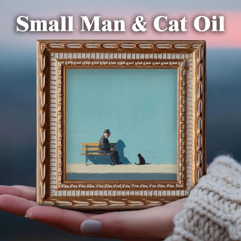 May include: A framed oil painting titled "Small Man & Cat Oil" depicts a man seated on a bench with a black cat. The artwork features a light blue background and a gold-colored ornate frame. The painting is held in a person's hand.