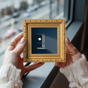May include: A small, square, gold-colored framed art piece featuring a minimalist design of a window with a circular element. The artwork is set against a dark blue background. The frame is held by a person.