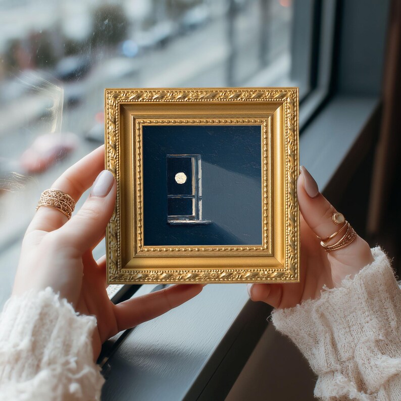 May include: A small, square, gold-colored framed art piece featuring a minimalist design of a window with a circular element. The artwork is set against a dark blue background. The frame is held by a person.