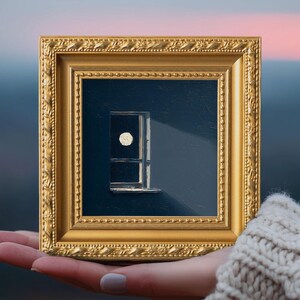 May include: A small framed painting depicts a window with a bright moon and a beam of light against a dark blue background. The painting is in a gold frame and held in a hand.