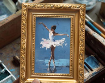 May include: A framed painting of a ballerina in a white tutu and pointe shoes, arms outstretched. The artwork is set against a blue background, with a gold ornate frame. The painting captures a moment of graceful dance.