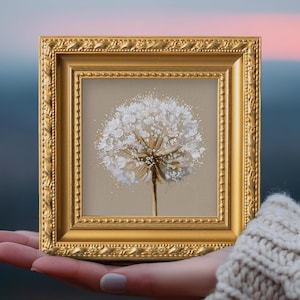May include: A square painting of a white dandelion seed head with textured brushstrokes on a light beige background, encased in an ornate, metallic gold frame with a beaded and rope-like decorative border detail along the edges.