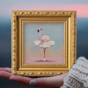 Op de afbeelding: Een ingelijst schilderij van een flamingo in een balletpakje en spitsschoenen. Het kunstwerk toont een roze flamingo met een zwarte snavel en een witte tutu. De lijst is goudkleurig en vierkant, met een gedetailleerde rand.