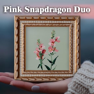 May include: A square painting titled Pink Snapdragon Duo featuring two pink snapdragon flowers with green stems on a light sage green background, encased in an ornate gold frame with a textured, repeating leaf pattern along the border.