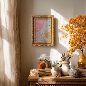May include: A framed abstract painting with a gold ornate frame. The artwork depicts a beach scene with figures walking along the shore. The painting has a pink, white, and yellow color palette. The painting is displayed on a wooden table with decorative items.