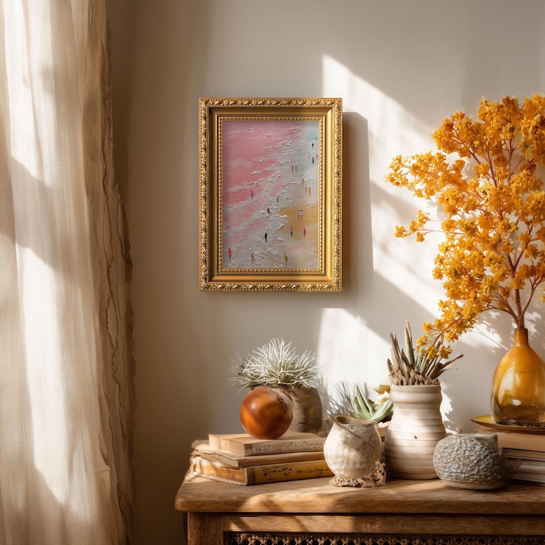 May include: A framed abstract painting with a gold ornate frame. The artwork depicts a beach scene with figures walking along the shore. The painting has a pink, white, and yellow color palette. The painting is displayed on a wooden table with decorative items.