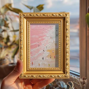 May include: A small framed painting with a gold ornate frame. The artwork depicts a beach scene with a pink and white sky and a yellow beach. Tiny figures of people are walking on the beach. The painting is held by a person.