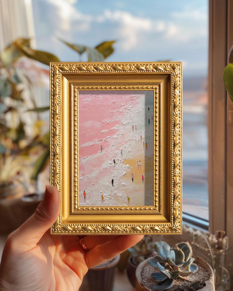 May include: A small framed painting with a gold ornate frame. The artwork depicts a beach scene with a pink and white sky and a yellow beach. Tiny figures of people are walking on the beach. The painting is held by a person.