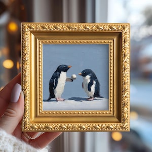 以下が含まれることがあります： 2羽のペンギンを描いた、小さく正方形の額装された絵画。1羽のペンギンが小さな白いボールを持っており、もう1羽のペンギンはそれを見ています。絵画は水色の背景で、金色のフレームに入っています。フレームは約15cmです。