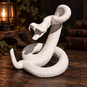 Puede incluir: Una figura de serpiente de cascabel blanca en una pose enroscada y de ataque con la boca abierta, mostrando los colmillos. La escultura presenta un patrón de escamas texturizado y muy detallado en todo el cuerpo, con la cola levantada.
