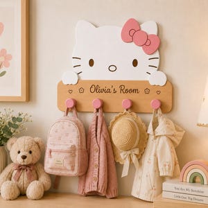 Può includere: Un appendiabiti da parete Hello Kitty bianco con un fiocco rosa, caratterizzato da un pannello orizzontale in legno chiaro inciso con il testo 'Olivia's Room' e quattro ganci circolari rosa che reggono uno zainetto, un cardigan e un cappello.