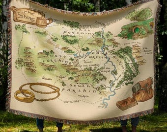 Może przedstawiać: Beżowy koc z mapą Shire z Władcy Pierścieni. Mapa zawiera ilustracje hobbitich dziur, pierścieni i tekstu. Koc ma frędzel i jest prezentowany na zewnątrz.
