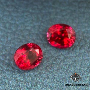 Peut inclure: Deux pierres précieuses rouges vives de forme ovale sont présentées sur un fond texturé de couleur sarcelle. Les gemmes ont de multiples facettes, reflétant la lumière et créant un effet scintillant. Le logo Ideal Gems Lab est visible en bas à droite.