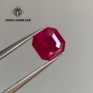 以下が含まれることがあります： 金属製のピンセットで保持された、八角形のファセットカットが施された深い赤色の宝石。左上隅には「IDEAL GEMS LAB」のロゴがあり、テキストの上にダイヤモンドのアイコンが表示されています。石は明確で鋭いエッジを備えています。