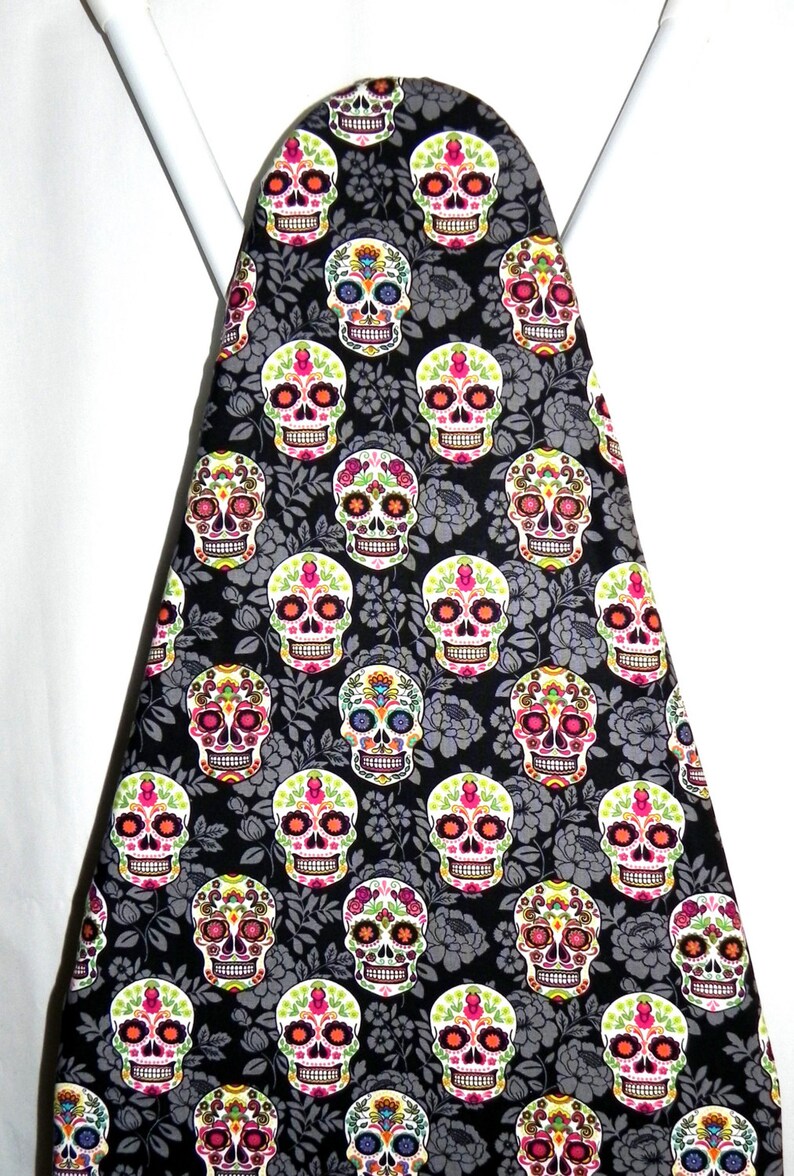 May include: An ironing board cover featuring a repeating pattern of colorful sugar skull designs on a black background. The skulls have intricate floral and geometric details in various colors, including pink, orange, and green.