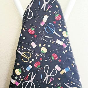 May include: Black ironing board cover with a repeating pattern of sewing tools and strawberries. The design includes scissors, spools of thread, needles, and buttons. The cover is mounted on a white ironing board frame.