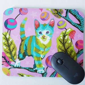 May include: A pink mouse pad with a colorful cat design. The cat is blue, green, and white with stripes. The background is pink with colorful circles and leaves.
