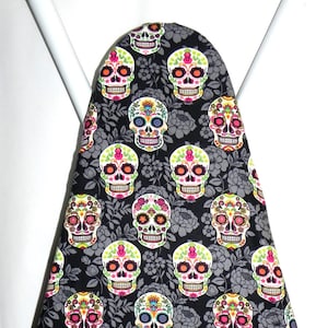 May include: An ironing board cover featuring a repeating pattern of colorful sugar skulls on a black background with gray floral accents. The skulls have vibrant pink, orange, green, and blue details.