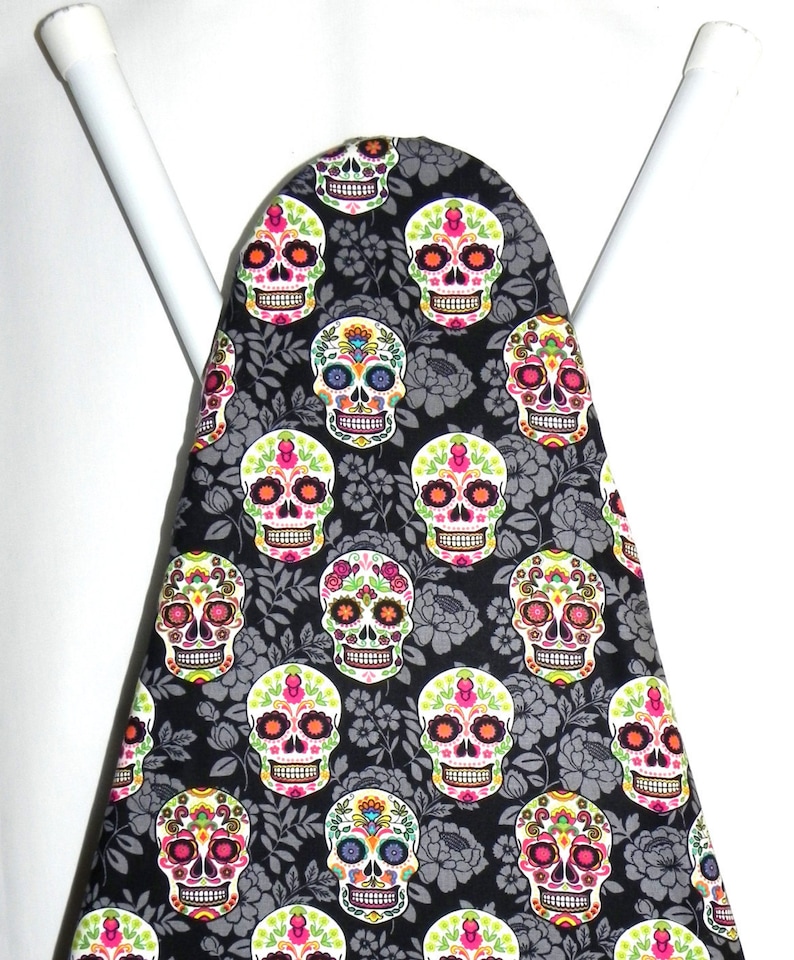 May include: An ironing board cover featuring a repeating pattern of colorful sugar skulls on a black background with gray floral accents. The skulls have vibrant pink, orange, green, and blue details.