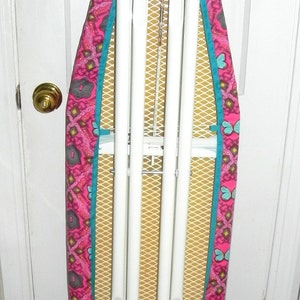 May include: A white metal ironing board with a pink, teal, and yellow butterfly pattern cover. The cover has a teal border and a yellow mesh ironing surface. The board is folded up against a white door.