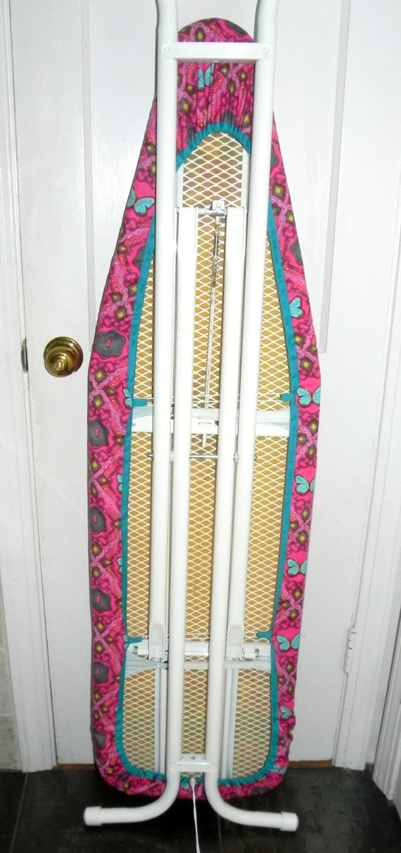 May include: A white metal ironing board with a pink, teal, and yellow butterfly pattern cover. The cover has a teal border and a yellow mesh ironing surface. The board is folded up against a white door.