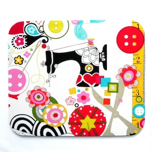 May include: A white mousepad with a colorful sewing-themed design. The design includes a black sewing machine, red heart, buttons, flowers, and a yellow measuring tape. The mousepad has rounded corners.