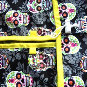 May include: A black fabric with a repeating pattern of colorful sugar skulls, flowers, and leaves. The fabric is trimmed with bright yellow ribbon and features a white toggle closure. The design is reminiscent of Dia de los Muertos.